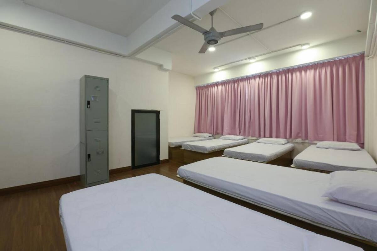 A room with four single beds, a grey metal wardrobe, a ceiling fan, and pink curtains.