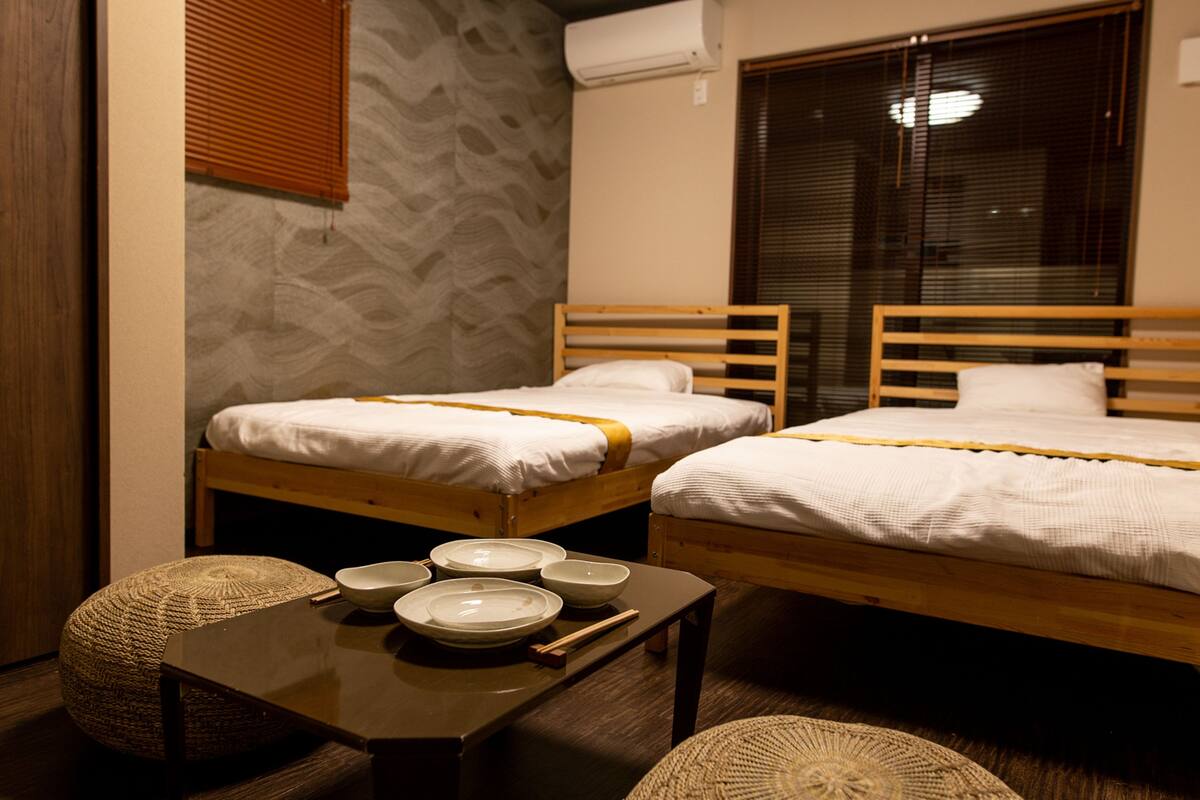A room with two wooden beds, a small table with bowls and chopsticks, and a woven pouf.