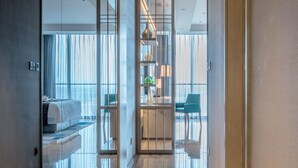 Corredor moderno de hotel com divisória de vidro, cama e área de estar com mesa e cadeiras.