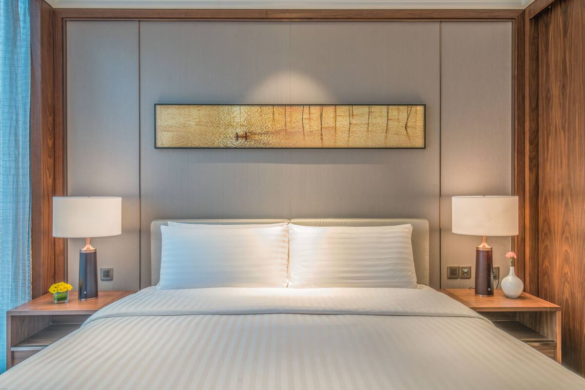 Una cama bien tendida con ropa blanca, dos mesitas de noche con lámparas, y un cuadro abstracto grande en la pared.