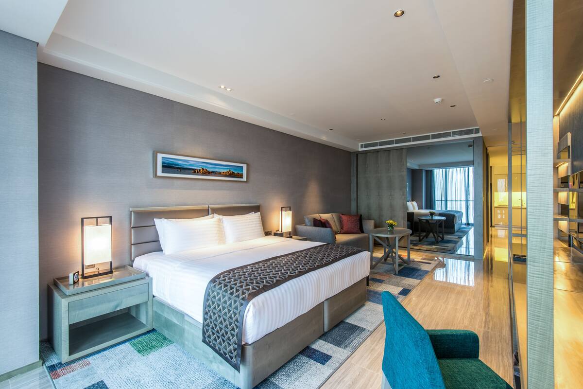 Una habitación de hotel moderna con una cama grande, mesitas de noche, una zona de estar con sofá y sillones, y vista a la habitación contigua.
