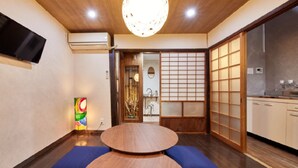 A traditional Japanese-style room with a tatami mat floor, a round wooden table, a television mounted on the wall, and a sliding door leading to a kitchen area. - Kyomachiya KANAU (Kyoto)