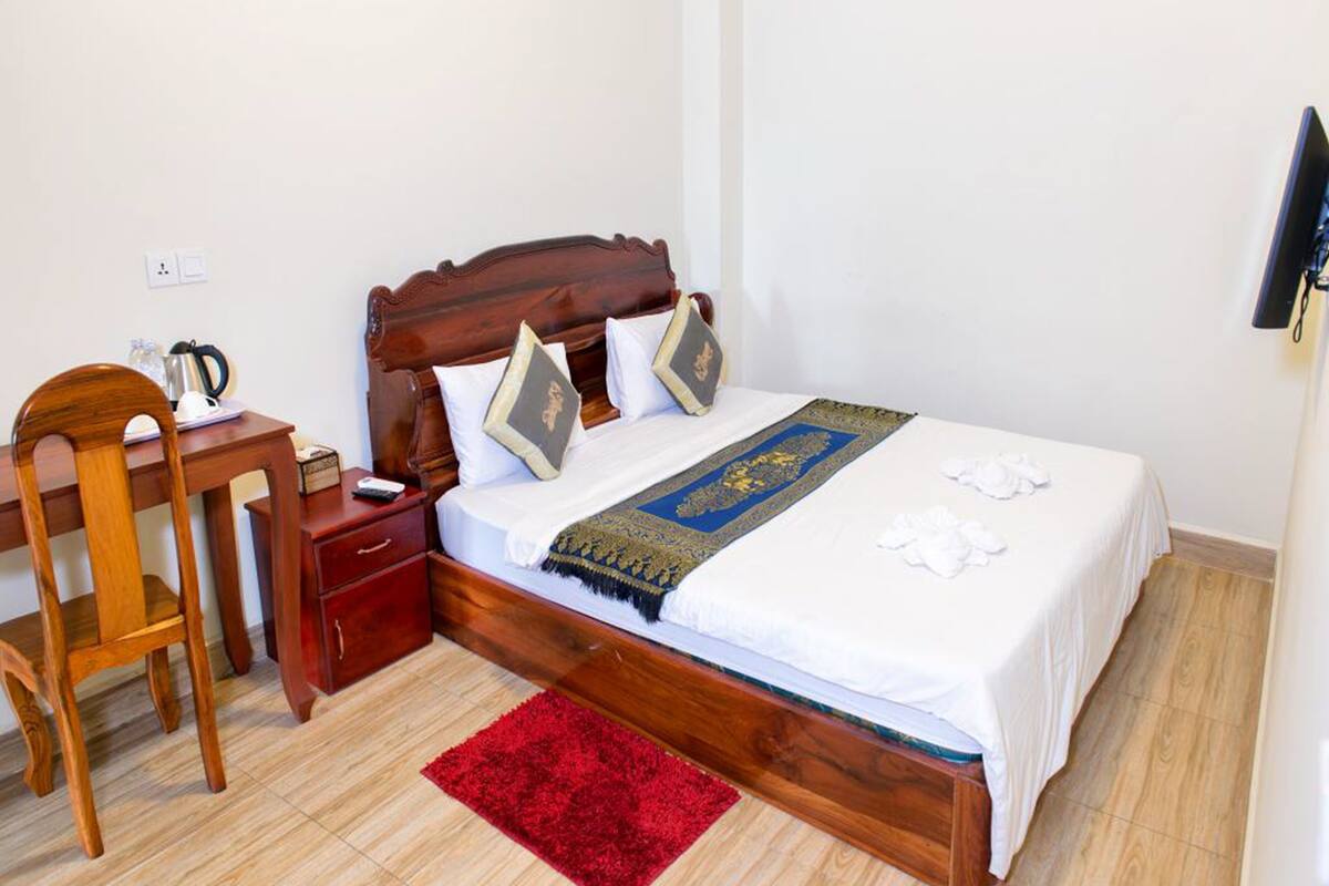 Een netjes opgemaakte slaapkamer met een houten bed, een rode vloerkleden, een houten stoel, een nachtkastje en een klein tafeltje met waterkoker en kopjes.