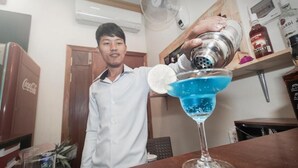 A person preparing a blue cocktail in a bar setting with a fan, shelves with bottles, and a wooden door in the background. - Dynsey Boutique Hotel (Phnom Penh)
