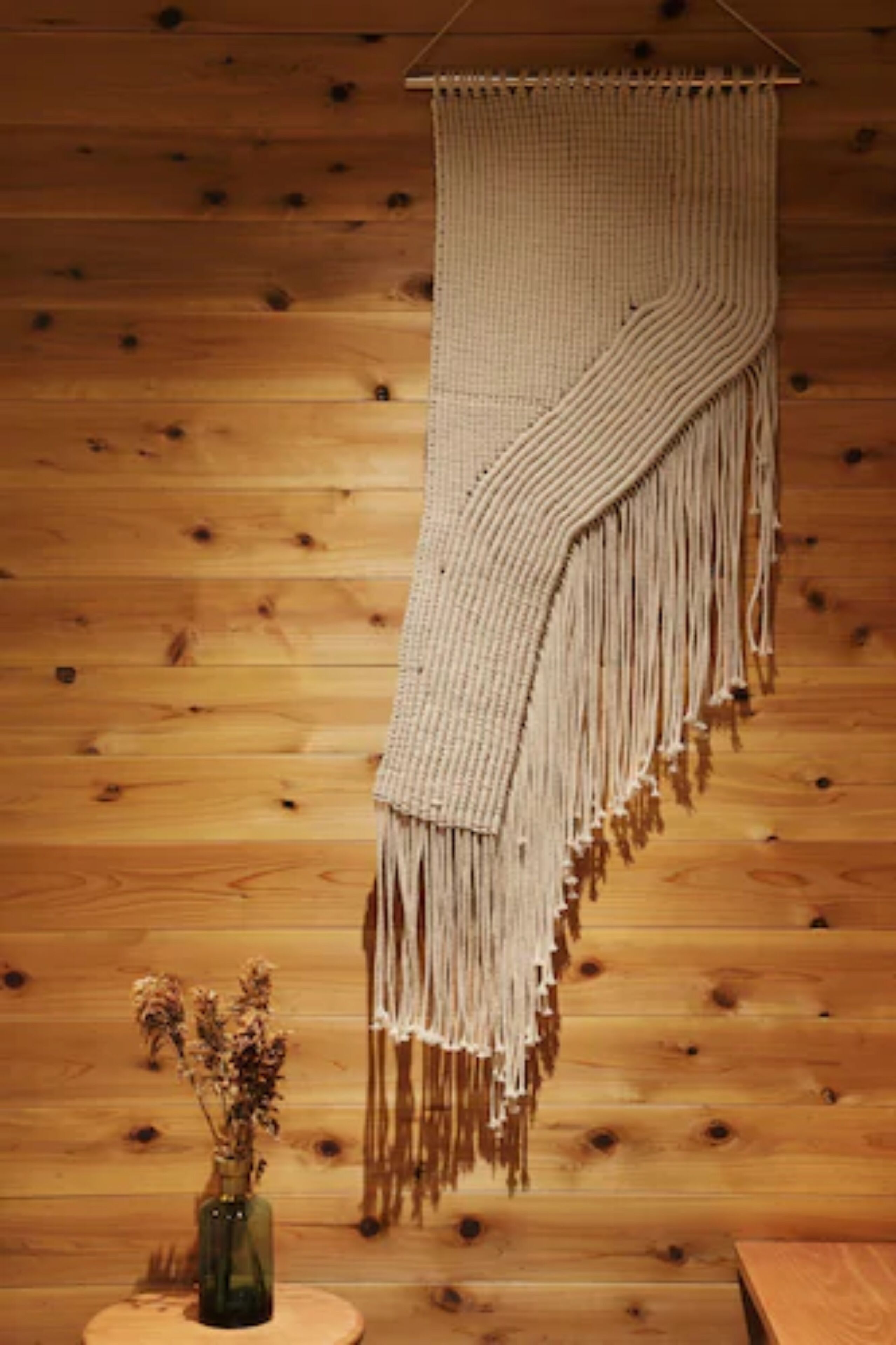 Eine Wanddeko aus Makramee mit Fransen, ein kleiner Holztisch mit einer Vase getrockneter Blumen und eine Holzwand.
