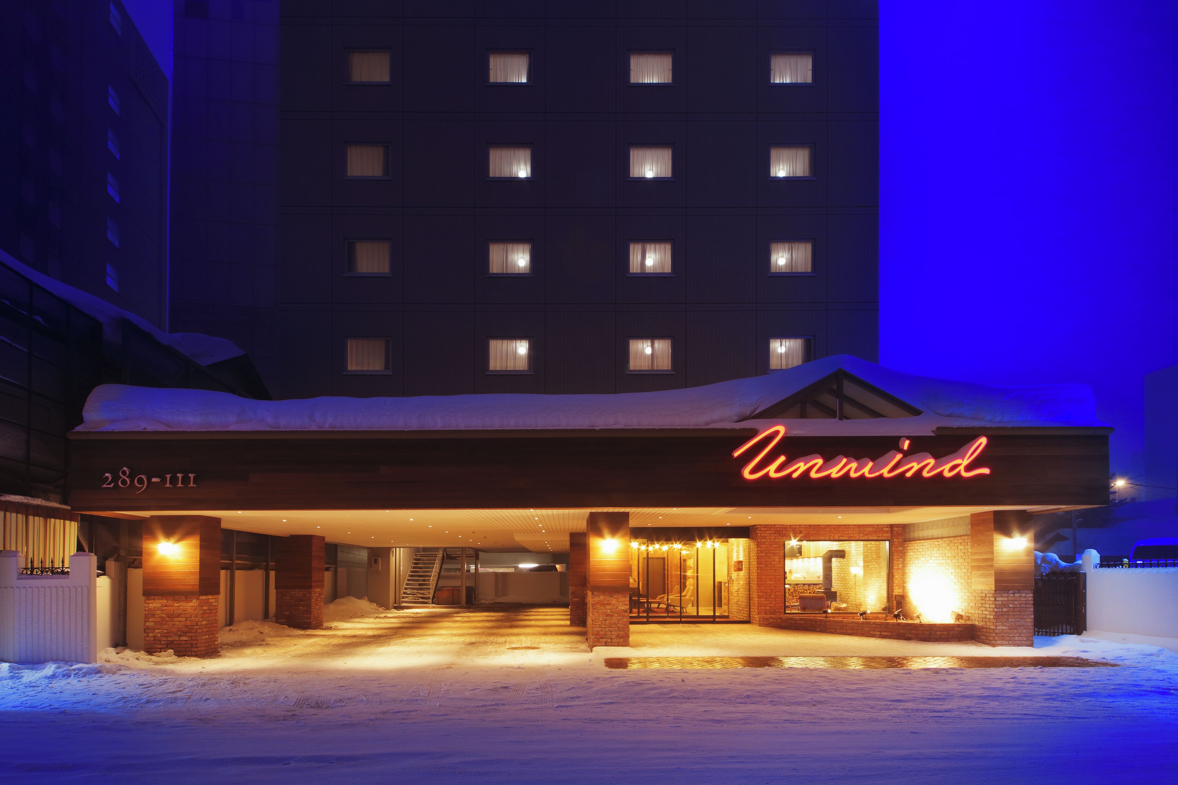 A hotel entrance with a neon sign, snow-covered ground, and a building in the background.