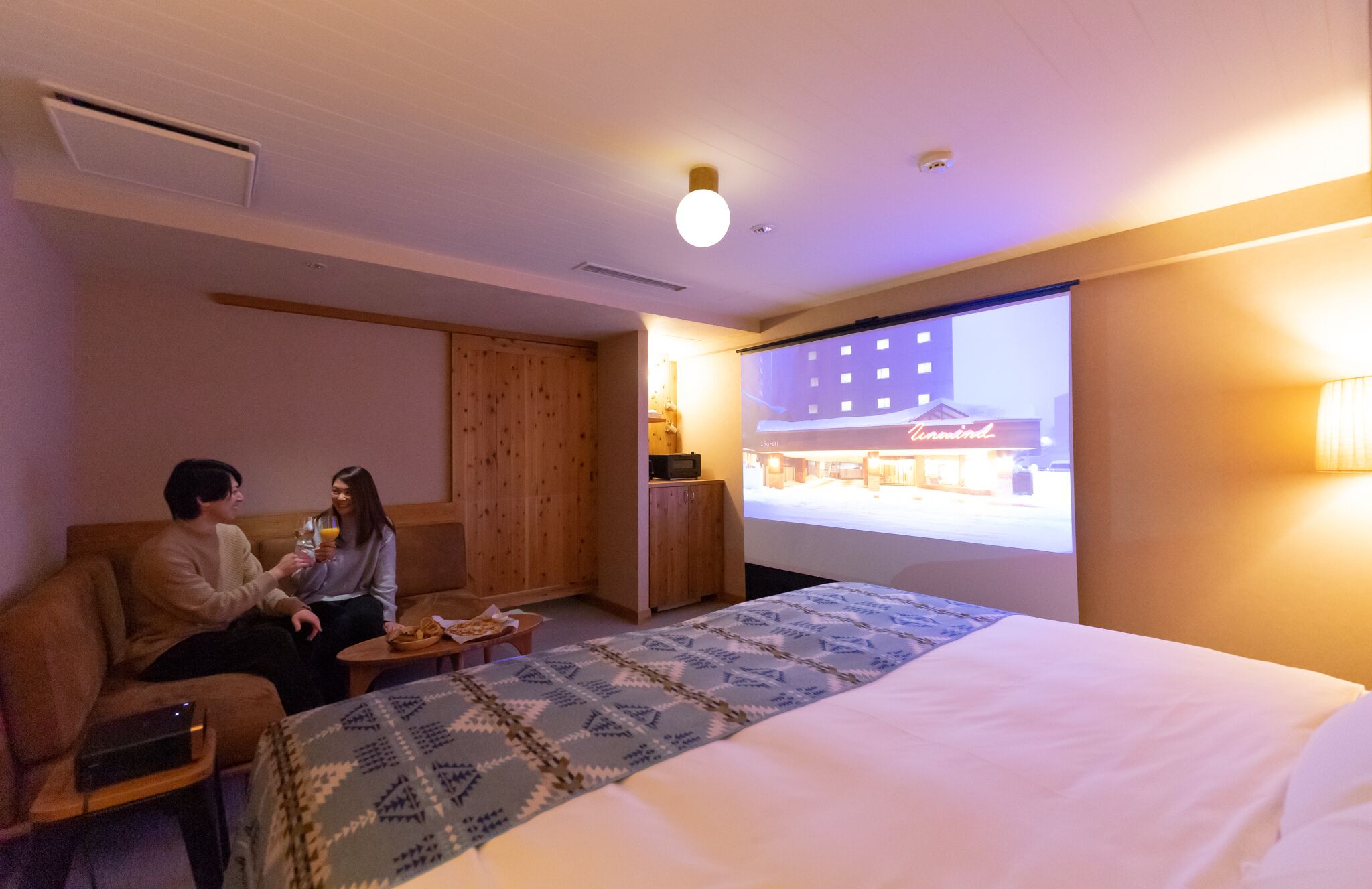 A hotel room with a bed, a sofa, a small table with snacks, a TV screen displaying a building, and a lamp.