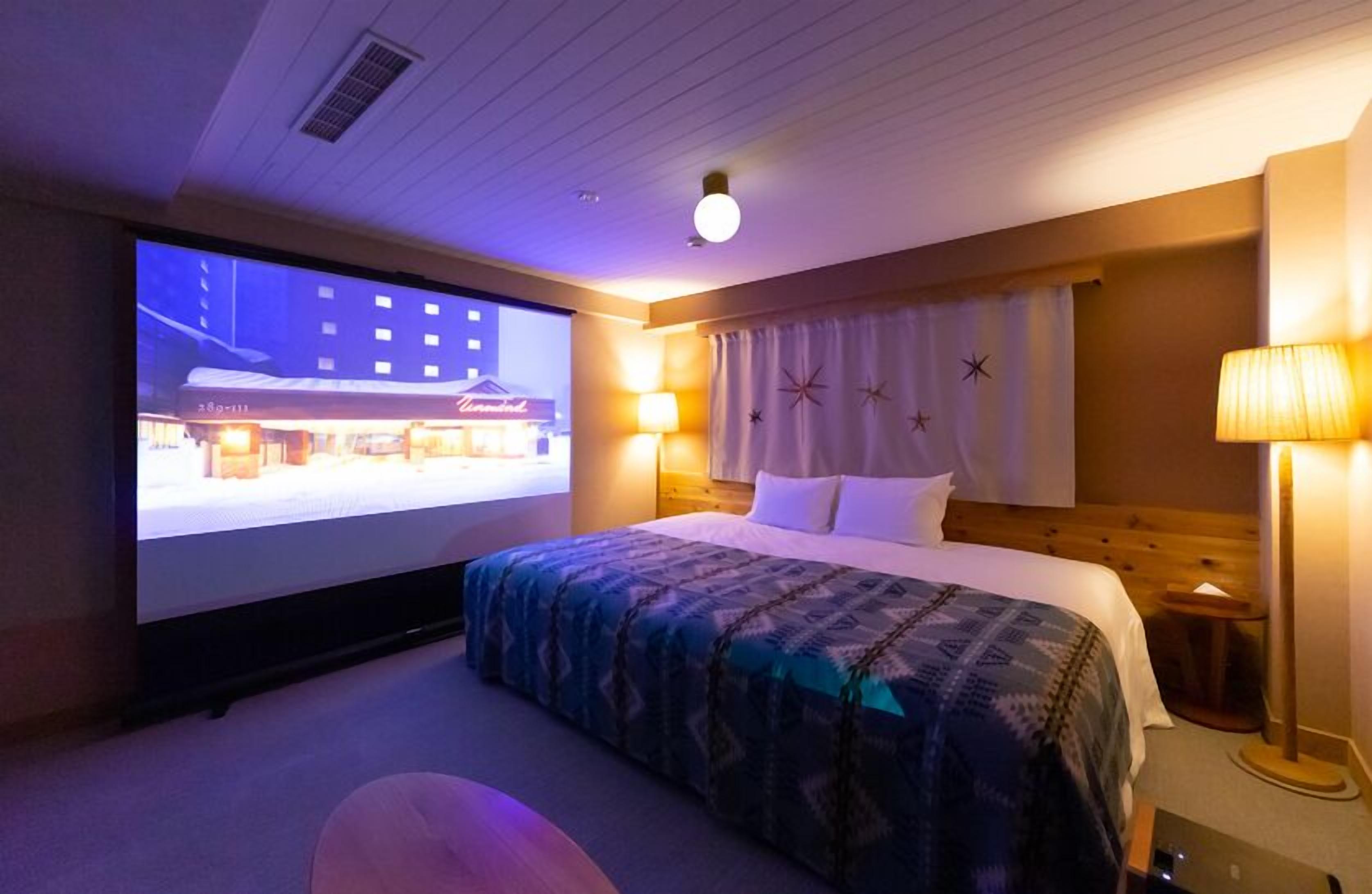 A hotel room with a bed, a projector screen, a lamp, and a nightstand.