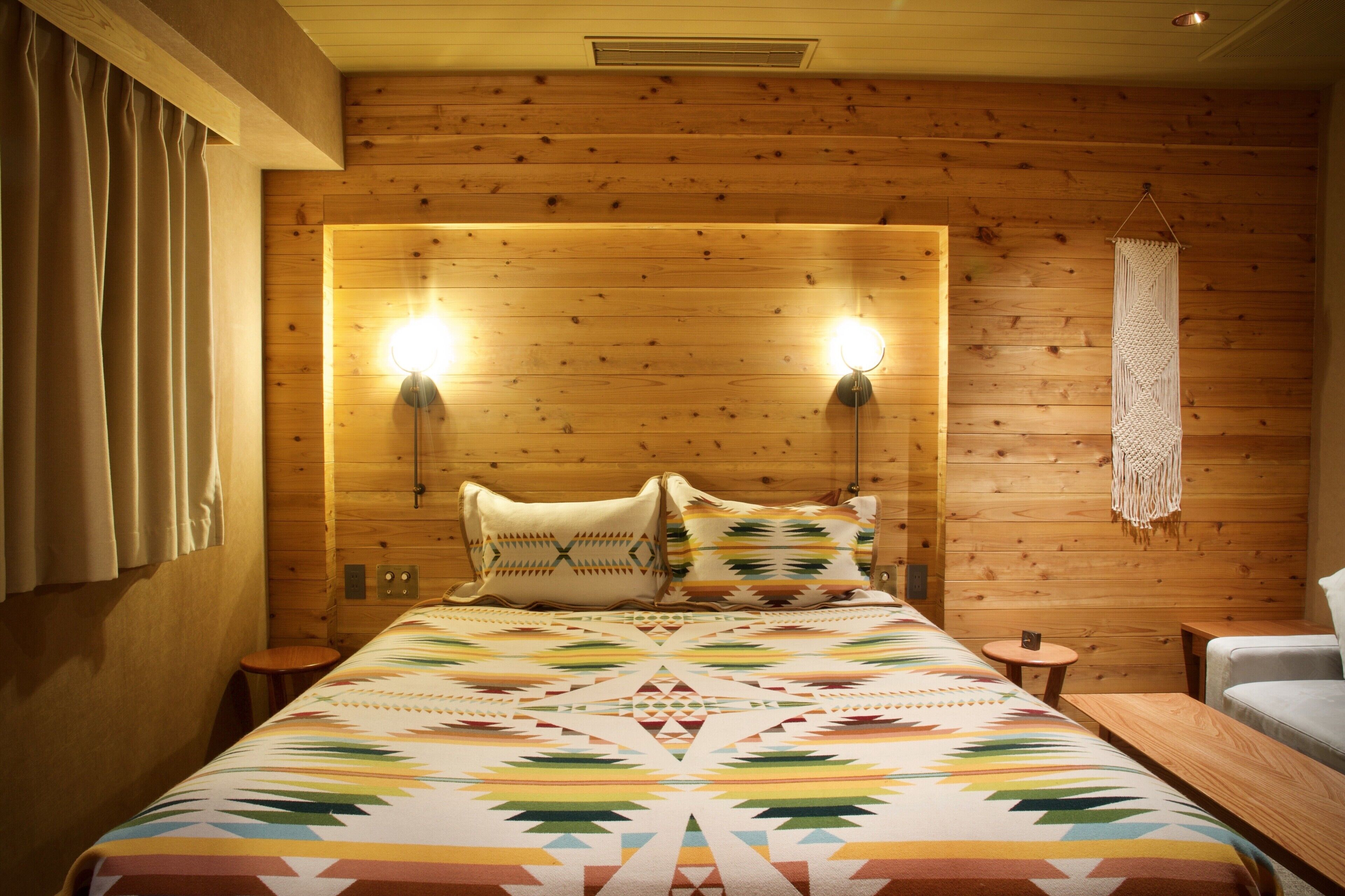 A wooden-paneled room with a bed, two wall-mounted lights, a macrame wall hanging, and a small round table.