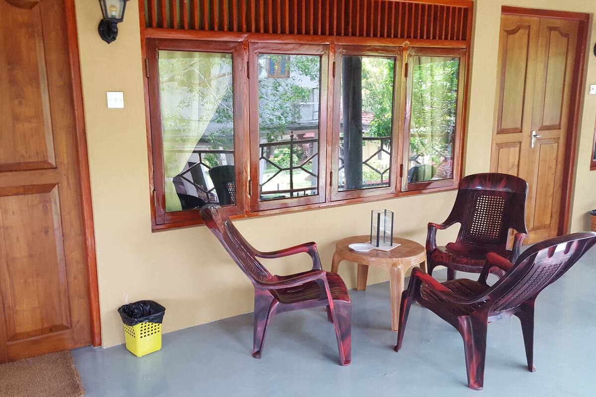 A room with wooden doors, a window with a view of greenery, two plastic chairs, a small table with a plate and a glass, and a yellow trash bin.