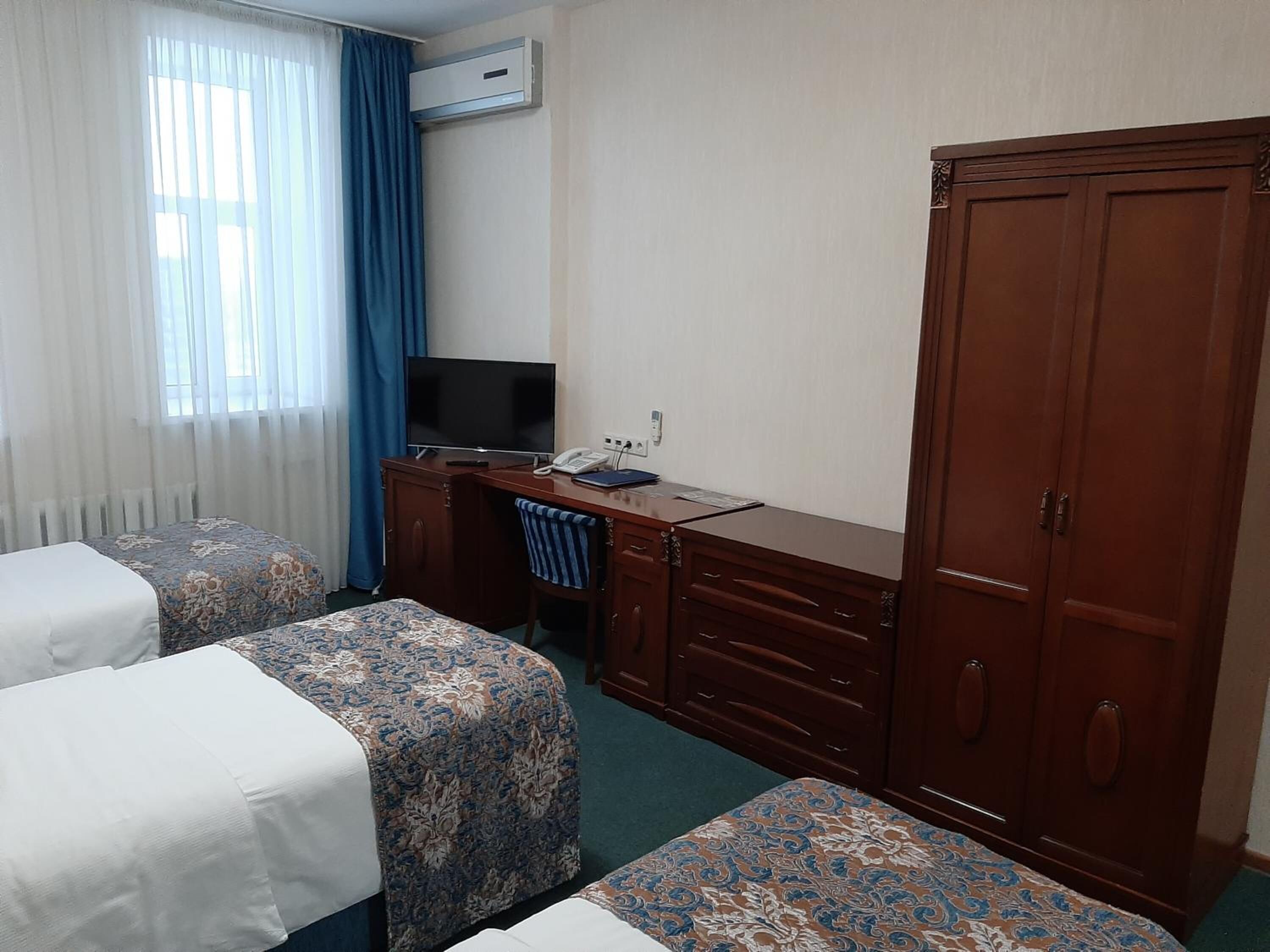 A hotel room with two beds, a desk with a TV, a wardrobe, and an air conditioning unit.