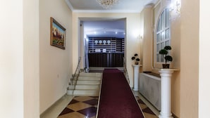Un couloir recouvert d’une moquette rouge menant à un escalier, avec des colonnes blanches et un lustre.