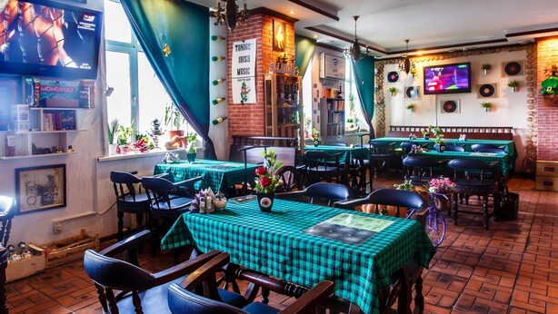 Un restaurant avec des tables à carreaux verts et des chaises noires, un mur en briques et un écran de télévision diffusant un film.