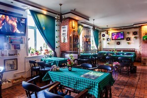 Um restaurante com mesas verde e branco quadriculadas e cadeiras pretas, parede de tijolos e uma tela de televisão exibindo um filme.