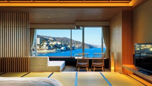 A hotel room with a large window offering a view of a coastal city, a bed, a TV, and a seating area with chairs. - Atami Korakuen Hotel (Atami)