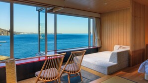 A room with a large window overlooking the sea, two wooden chairs, a sofa, and a tatami mat. - Atami Korakuen Hotel (Atami)