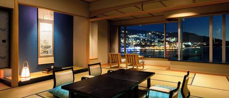 A traditional Japanese room with tatami flooring, a low table, and chairs, featuring a large window with a night view of a cityscape.