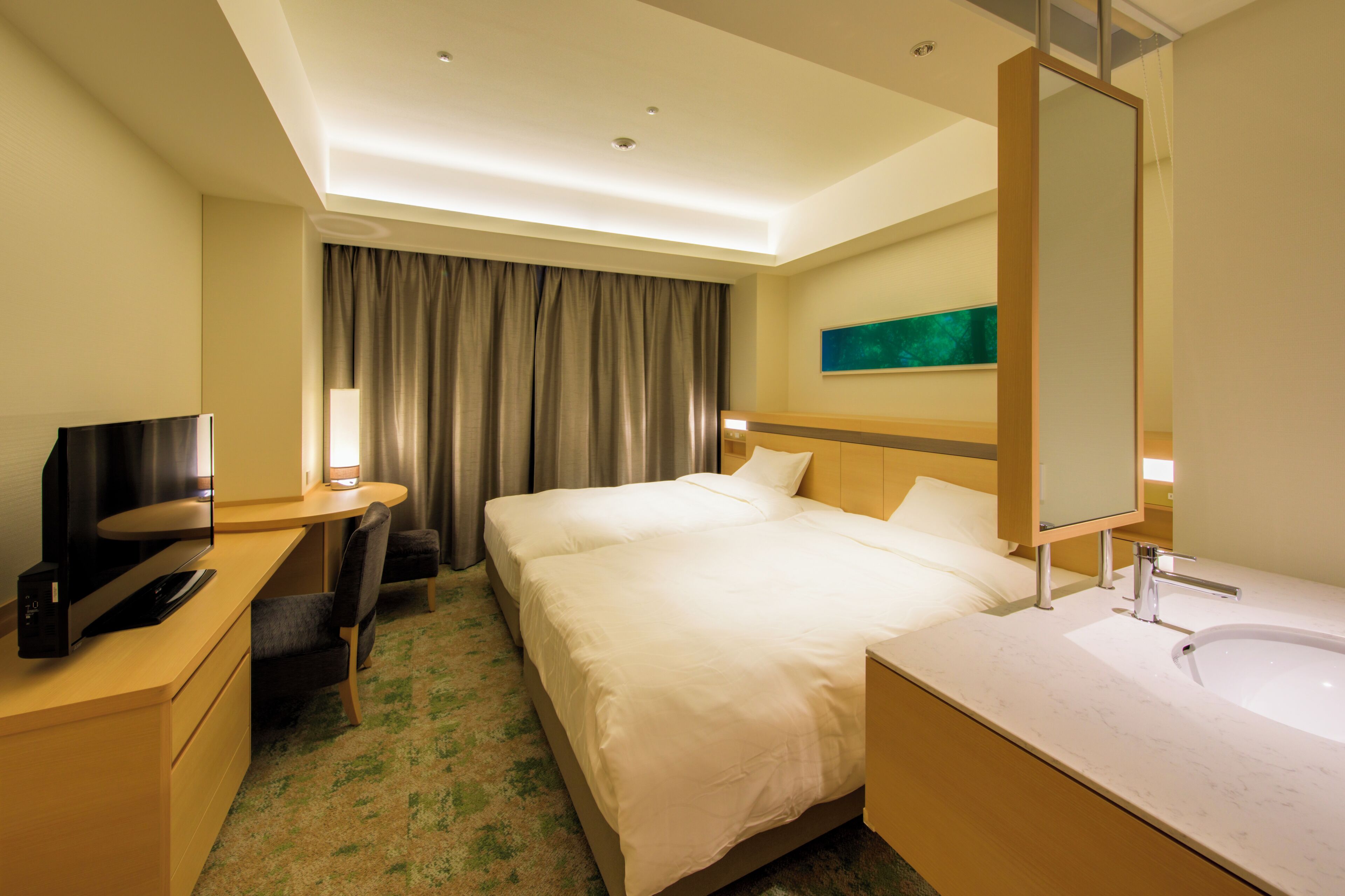 A hotel room with a bed, a desk with a computer, a TV, a large mirror, and a bathroom with a sink and toilet.