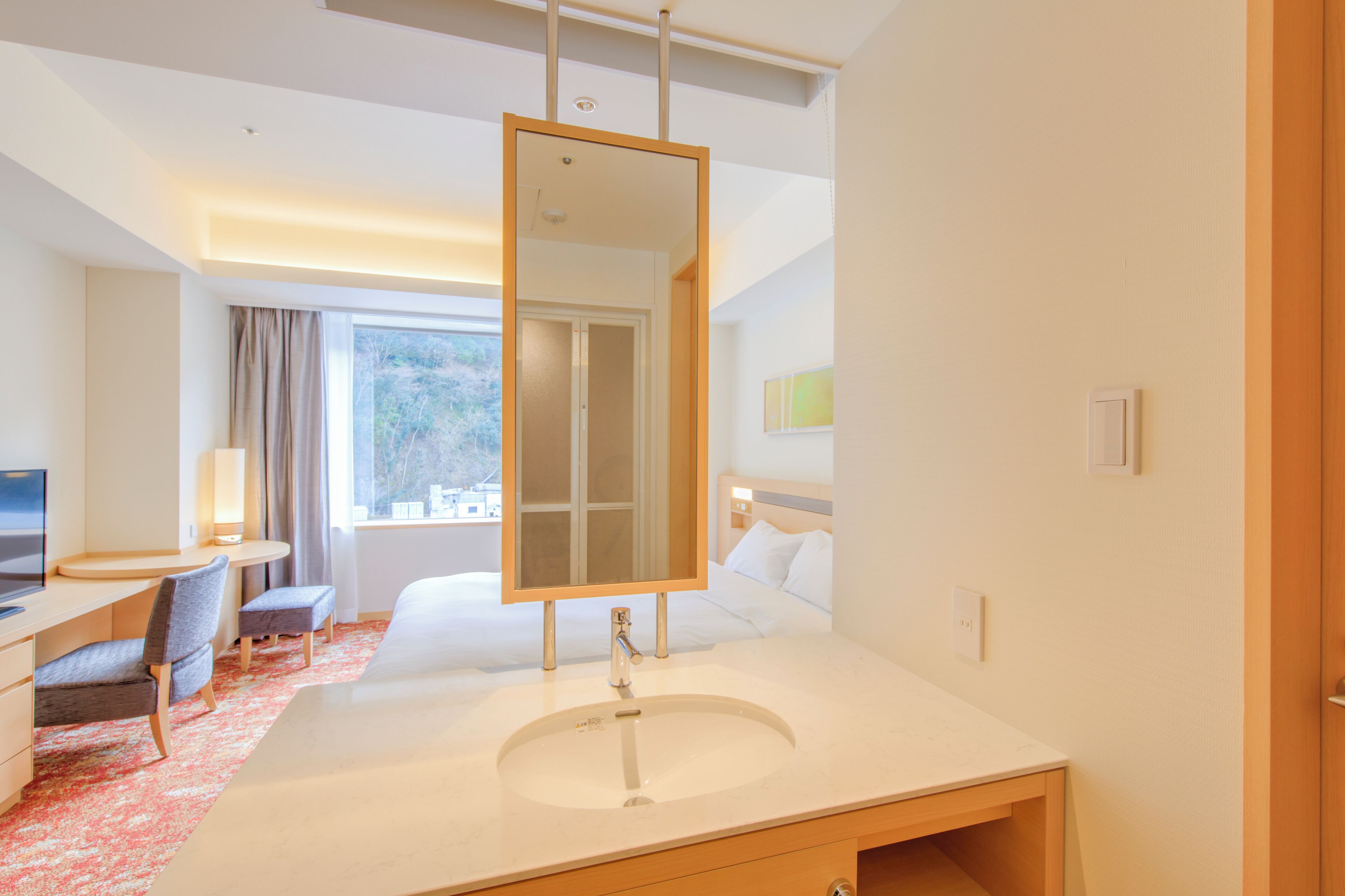 A hotel room with a large bed, a desk with a chair, a bathroom with a sink and mirror, and a window with curtains.