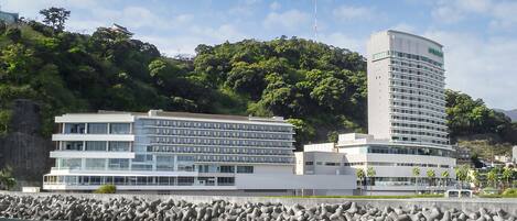 A coastal hotel with multiple floors, a rocky shoreline, and lush greenery in the background.