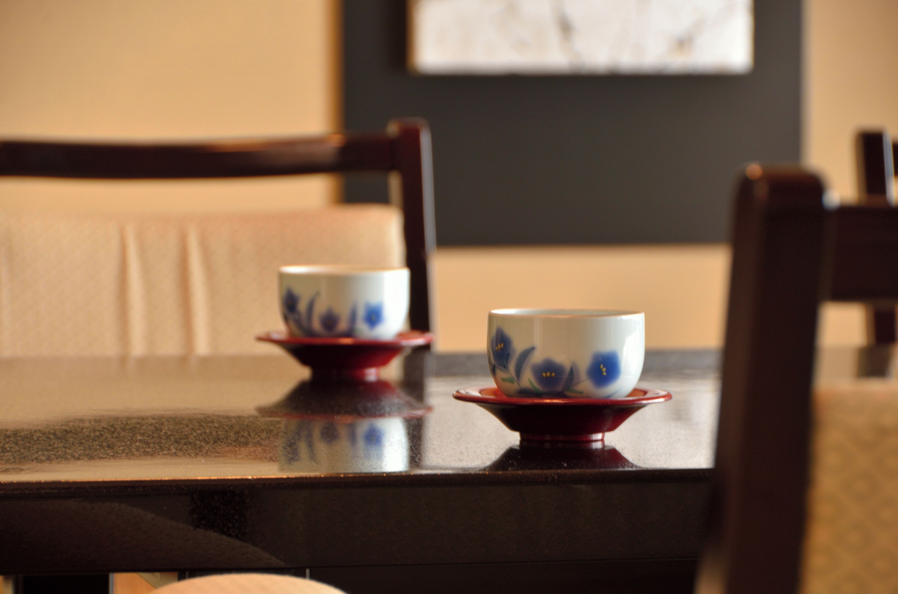 Une table à manger avec deux tasses en céramique posées sur des soucoupes, une chaise en bois et un tableau encadré accroché au mur.