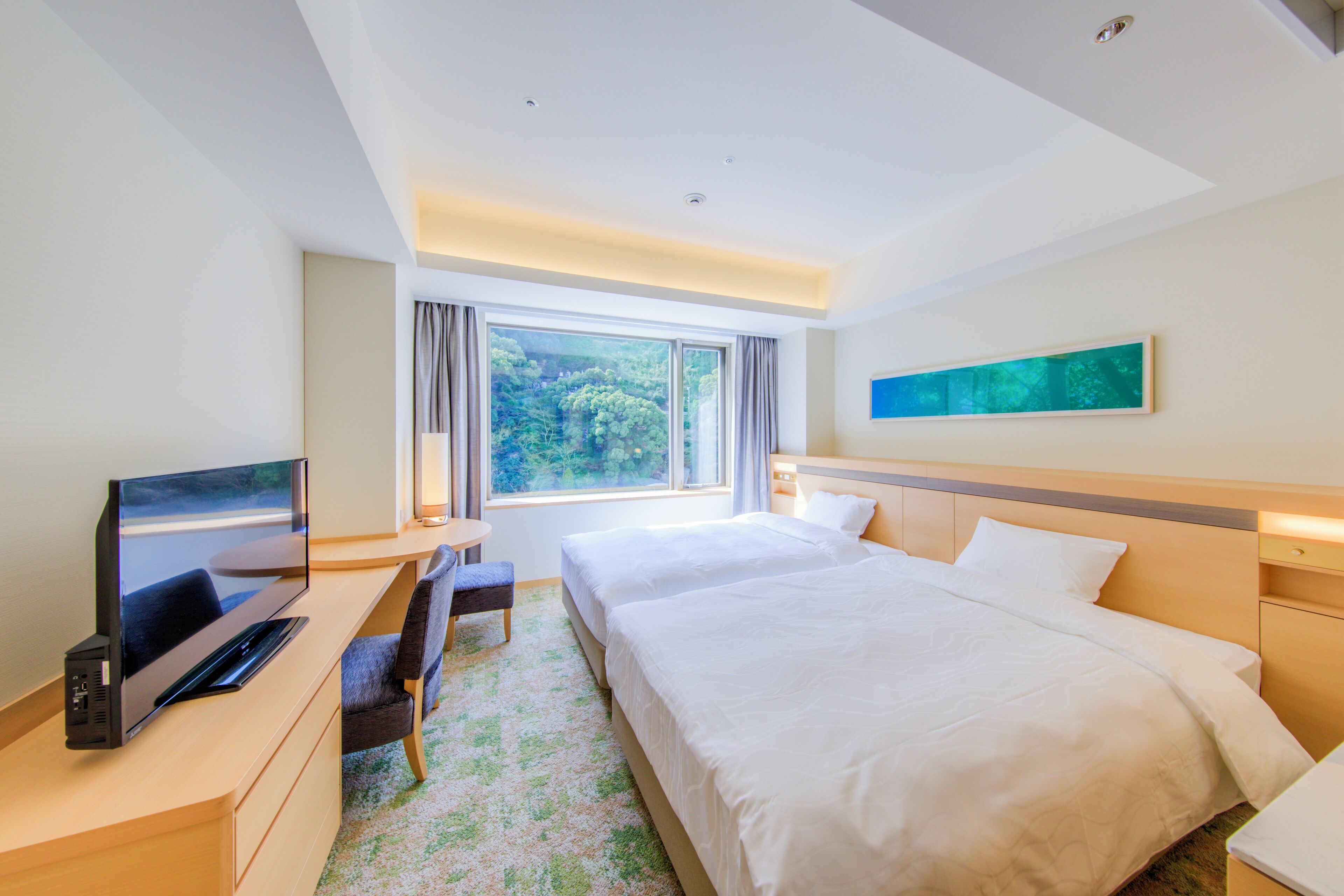 A hotel room with a bed, a desk with a flat-screen TV, a chair, and a window with a view of trees.