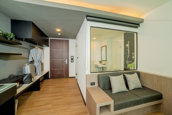 A modern hotel room with a wooden floor, a grey sofa, a wooden side table, a large mirror, and a wardrobe with a robe and bags. - Pause and Play Hotel (Chiang Mai)