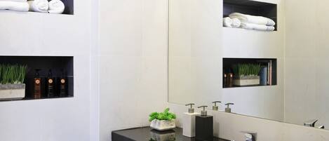 Un baño moderno con un inodoro blanco, un lavabo con espejo y estantes con toallas y artículos de aseo.