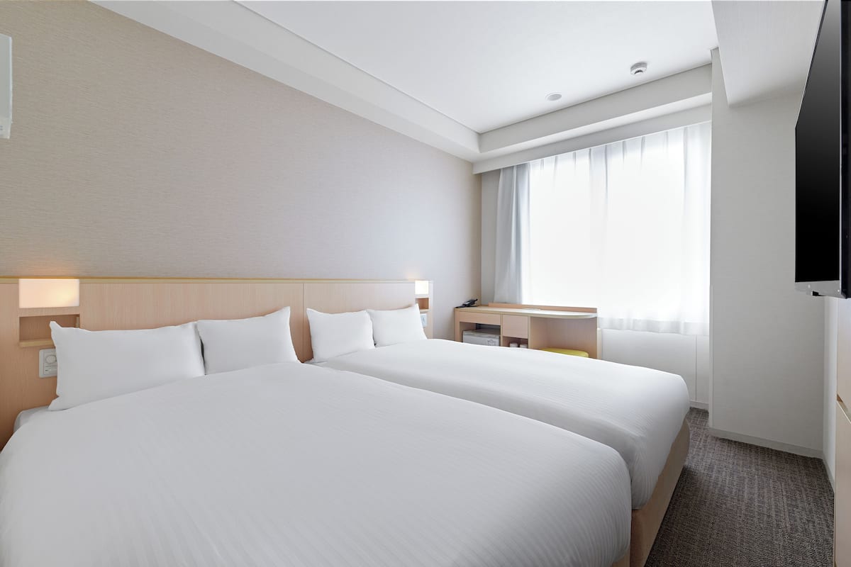Ein Hotelzimmer mit zwei Betten, einem Schreibtisch, einem Fernseher und einem großen Fenster mit Vorhängen.