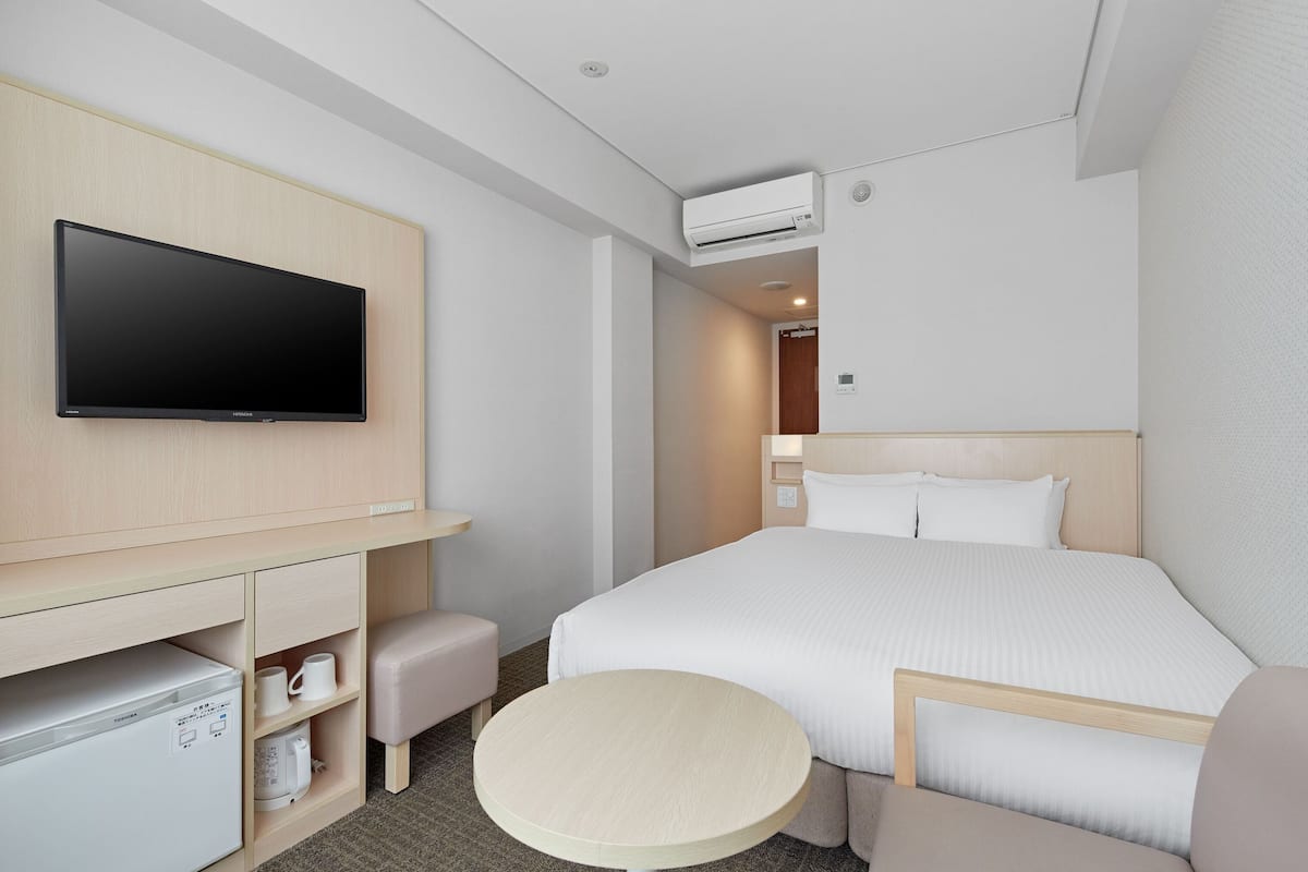 Ein Hotelzimmer mit Bett, Fernseher, Schreibtisch mit Computer, einem kleinen Tisch und einer Klimaanlage.