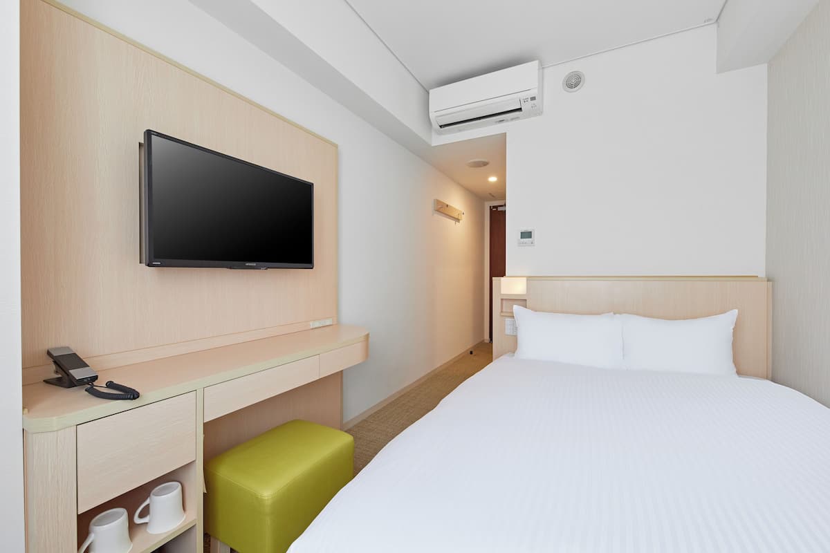 Ein Hotelzimmer mit einem Bett, einem Schreibtisch mit Flachbildfernseher, einem grünen Hocker und einer weißen Wand.