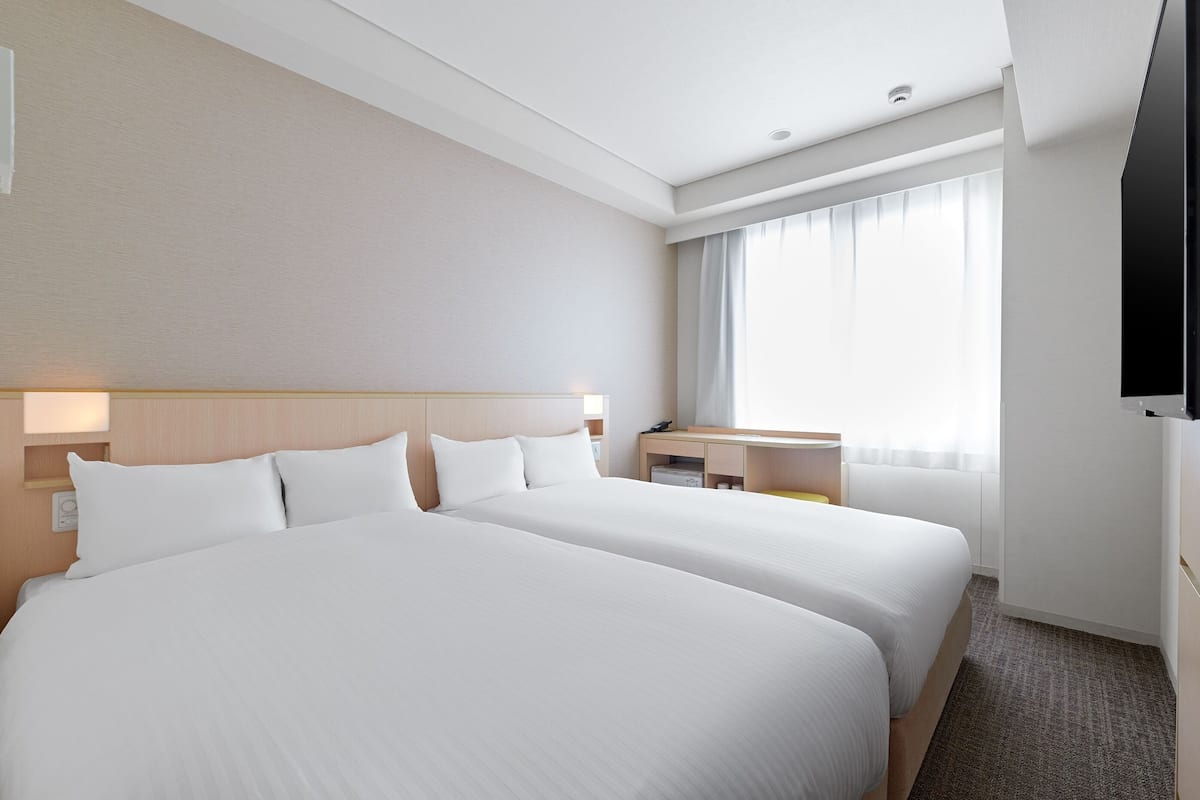Ein Hotelzimmer mit zwei Betten, einem Schreibtisch, einem Stuhl, einem Fernseher und einem Fenster mit Vorhängen.