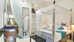 A four-poster canopy bed with a white bedspread and teal accents, a bedside table with a lamp, and a small bench in a room with a large mirror and a window with sheer curtains. - The Sun House (Galle)