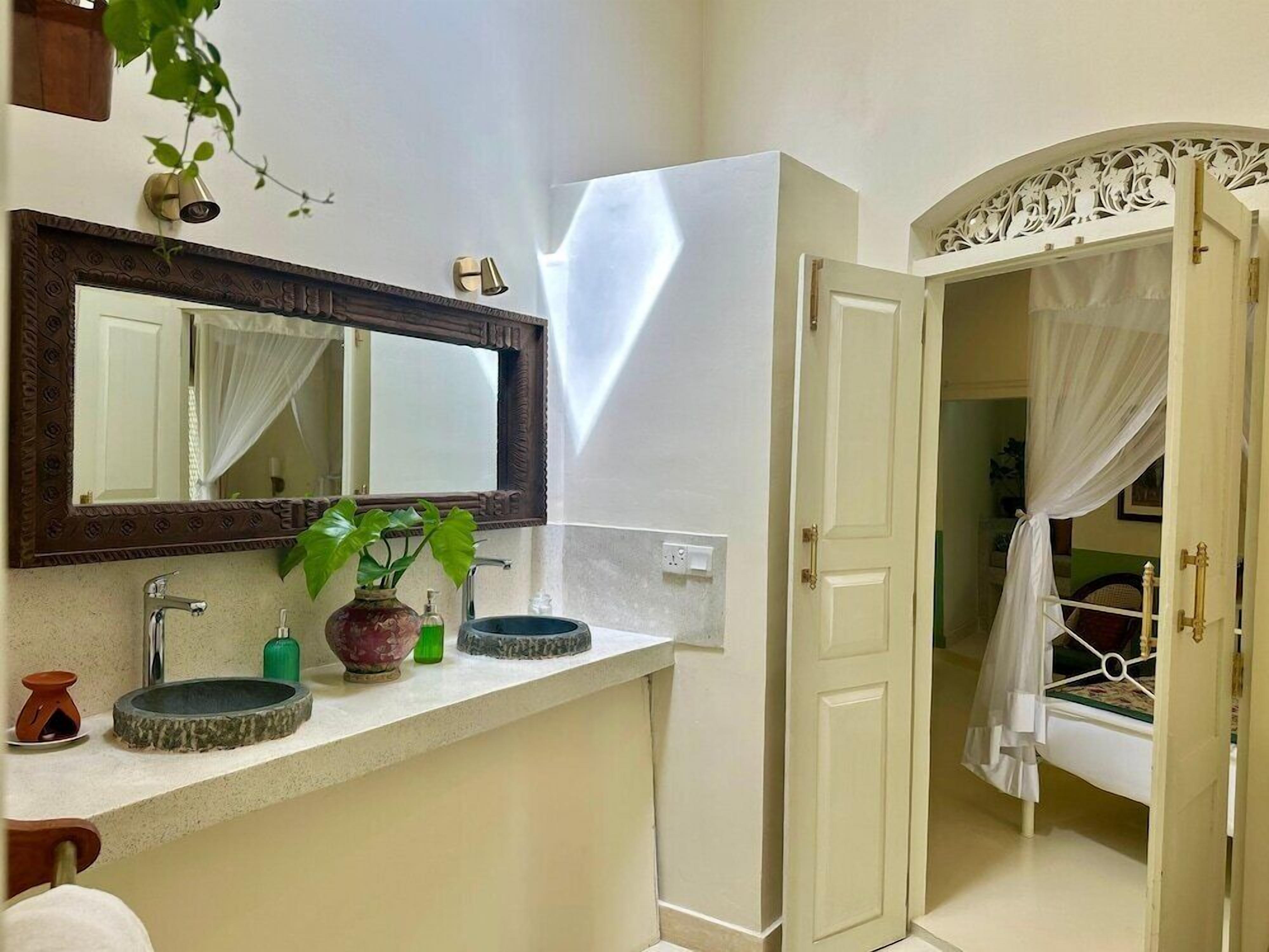 A bathroom with a large mirror, two sinks, a potted plant, and a door leading to a bedroom with a canopy bed.