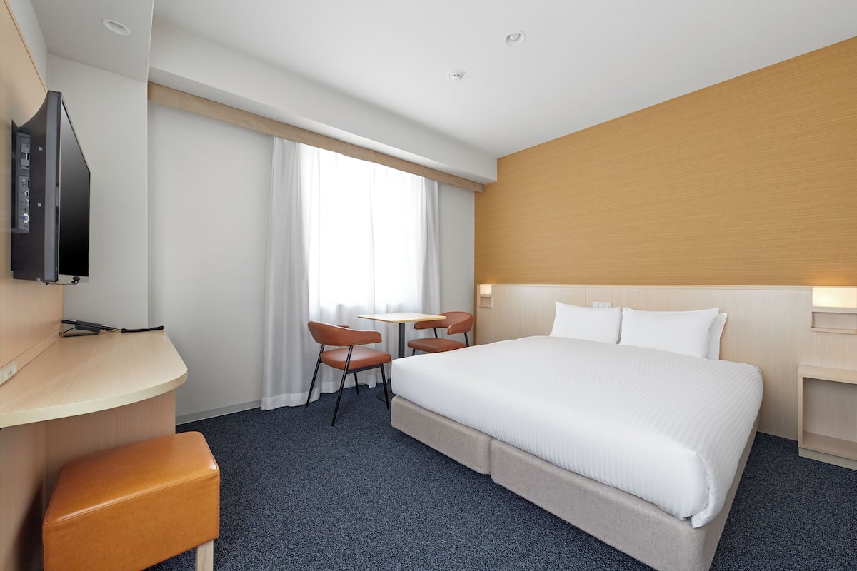 A hotel room with a bed, a desk, a TV, a small table, two chairs, and a window with curtains.