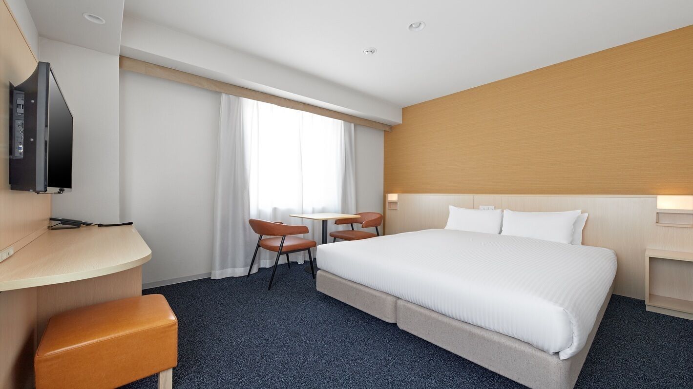Habitación de hotel con cama, escritorio, televisión, una pequeña mesa con sillas y ventana con cortinas.