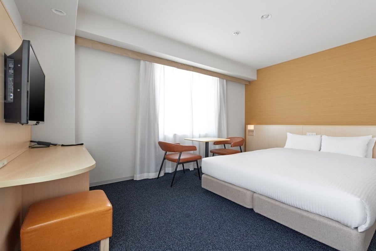 A hotel room with a bed, a desk, a TV, a small table with chairs, and a window with curtains.