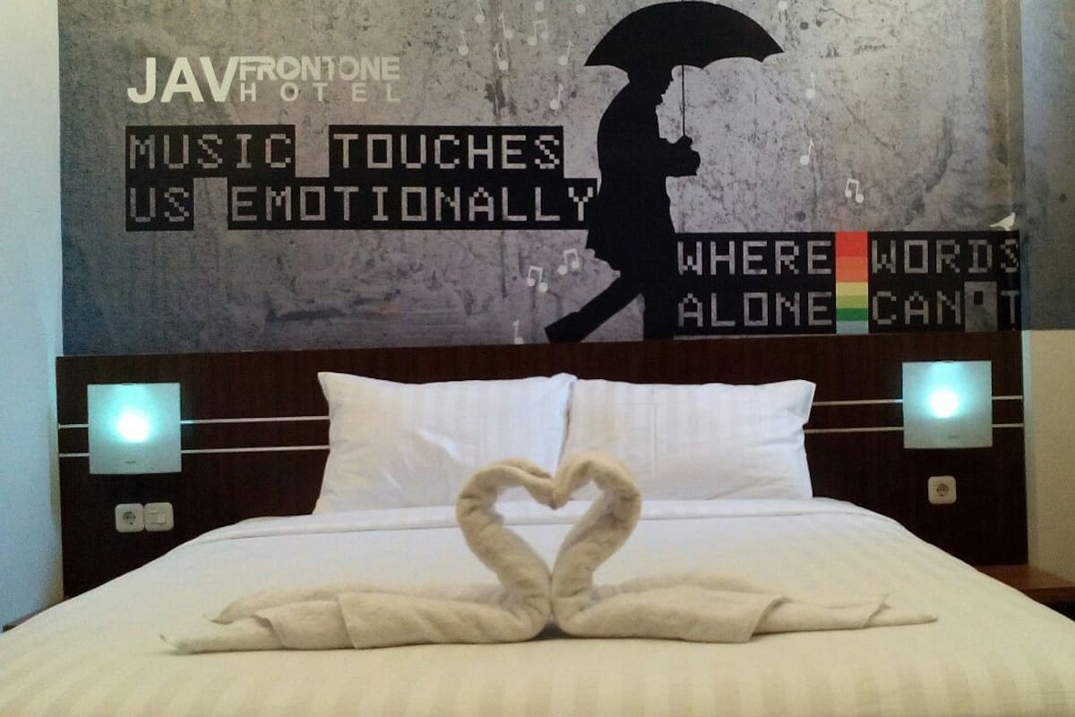 A hotel room with a bed featuring a heart-shaped towel arrangement, a wall with a silhouette of a person holding an umbrella, and text that reads "MUSIC TOUCHES US EMOTIONALLY WHERE WORDS ALONE CAN'T".