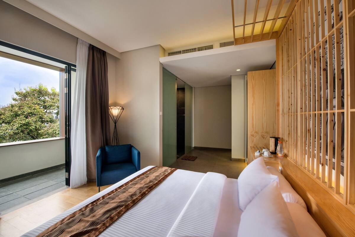 A modern hotel room with a large bed, a blue chair, a wooden wardrobe, and a view of trees through the window.