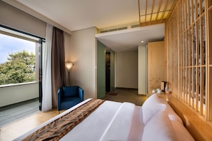 Habitación de hotel moderna con una cama grande, una silla azul, un armario de madera y vistas a árboles por la ventana.