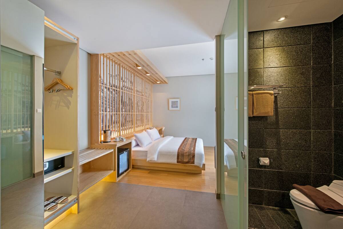 A modern hotel room with a bed, a small desk with a computer, a wooden wardrobe, and a bathroom with a dark tiled wall.