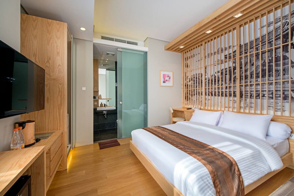 A modern hotel room with a wooden bed, a television, a desk, and a sliding door leading to a bathroom.