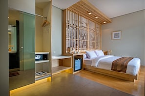 A modern hotel room with a wooden bed, a small desk, a built-in wardrobe, and a compact kitchenette.