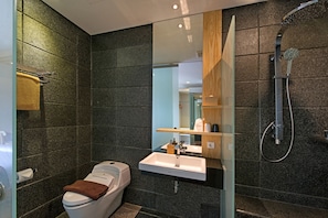 A modern bathroom with dark tiles, a white sink, a glass shower enclosure, and a wooden shelf with toiletries. - Tama Boutique Hotel (Bandung)
