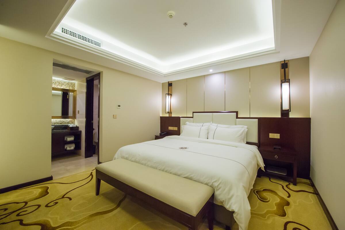 Une chambre d’hôtel avec un grand lit, une banquette, une table de chevet et une salle de bain visible à travers une porte ouverte.