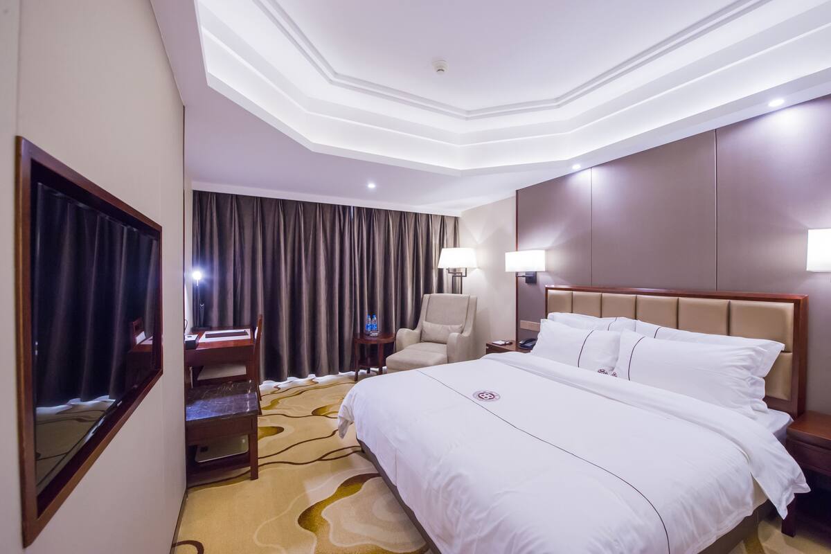 Une chambre d’hôtel avec un grand lit, un bureau avec une chaise, une petite table avec une lampe et une grande fenêtre avec des rideaux.