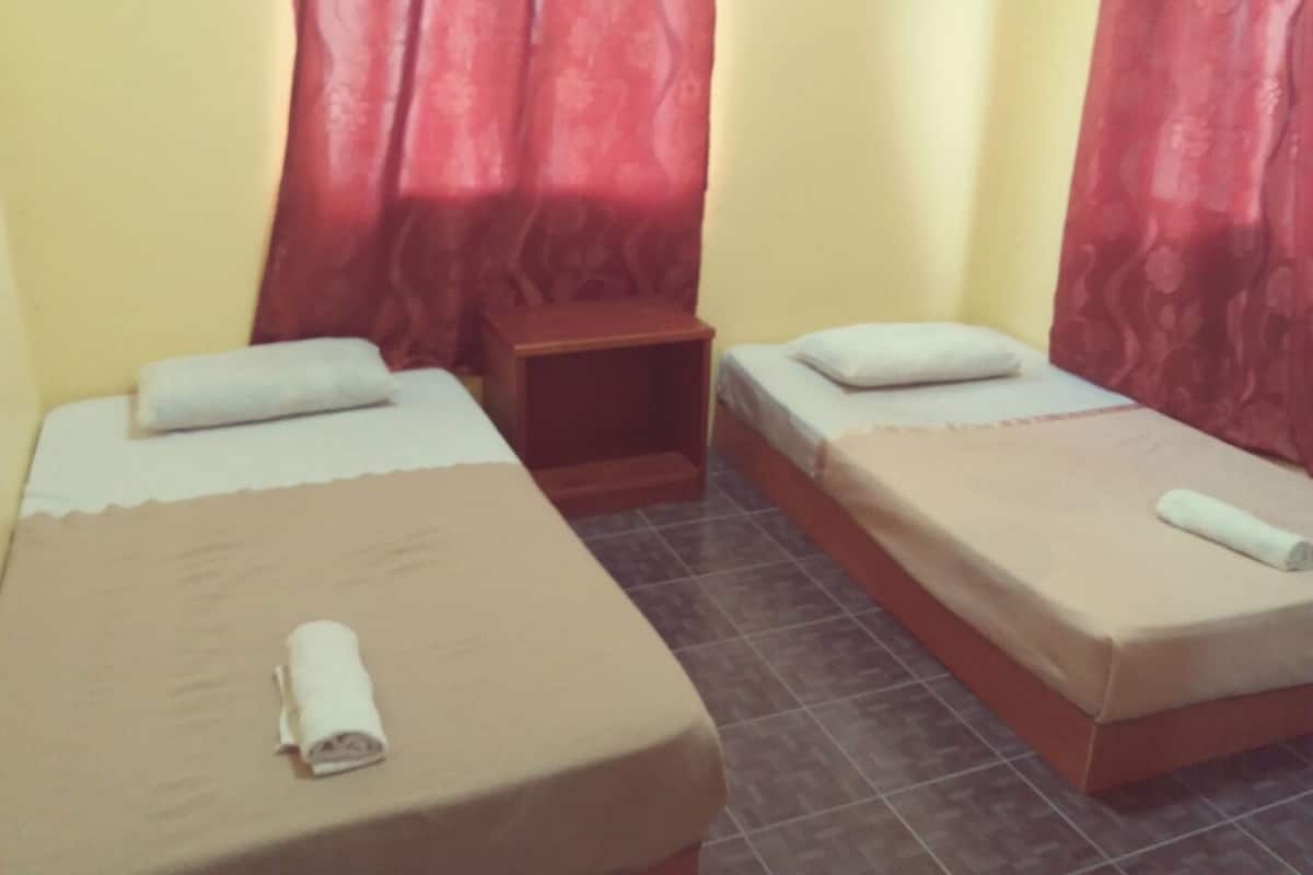 A room with two single beds, each with a pillow and a rolled towel. There is a small wooden nightstand between the beds.