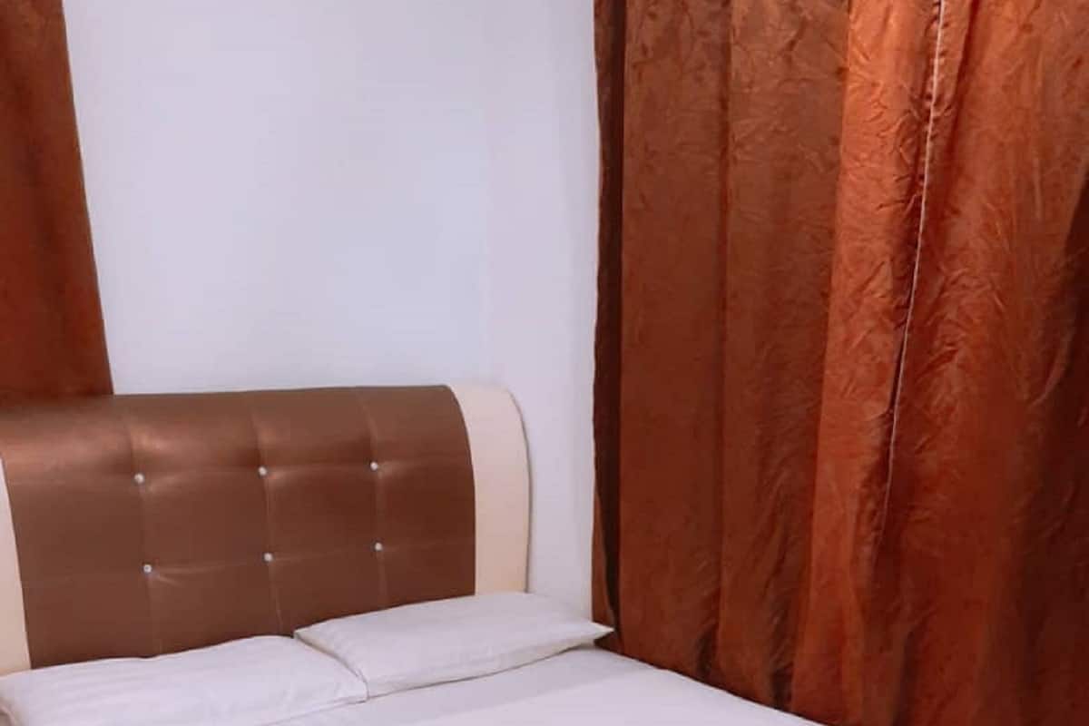 A bed with a brown headboard and white bedding in a room with a plain wall and a curtain.
