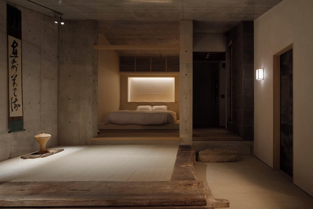 A minimalist bedroom with a bed, a bedside table, a wall-mounted light, and a window with blinds.