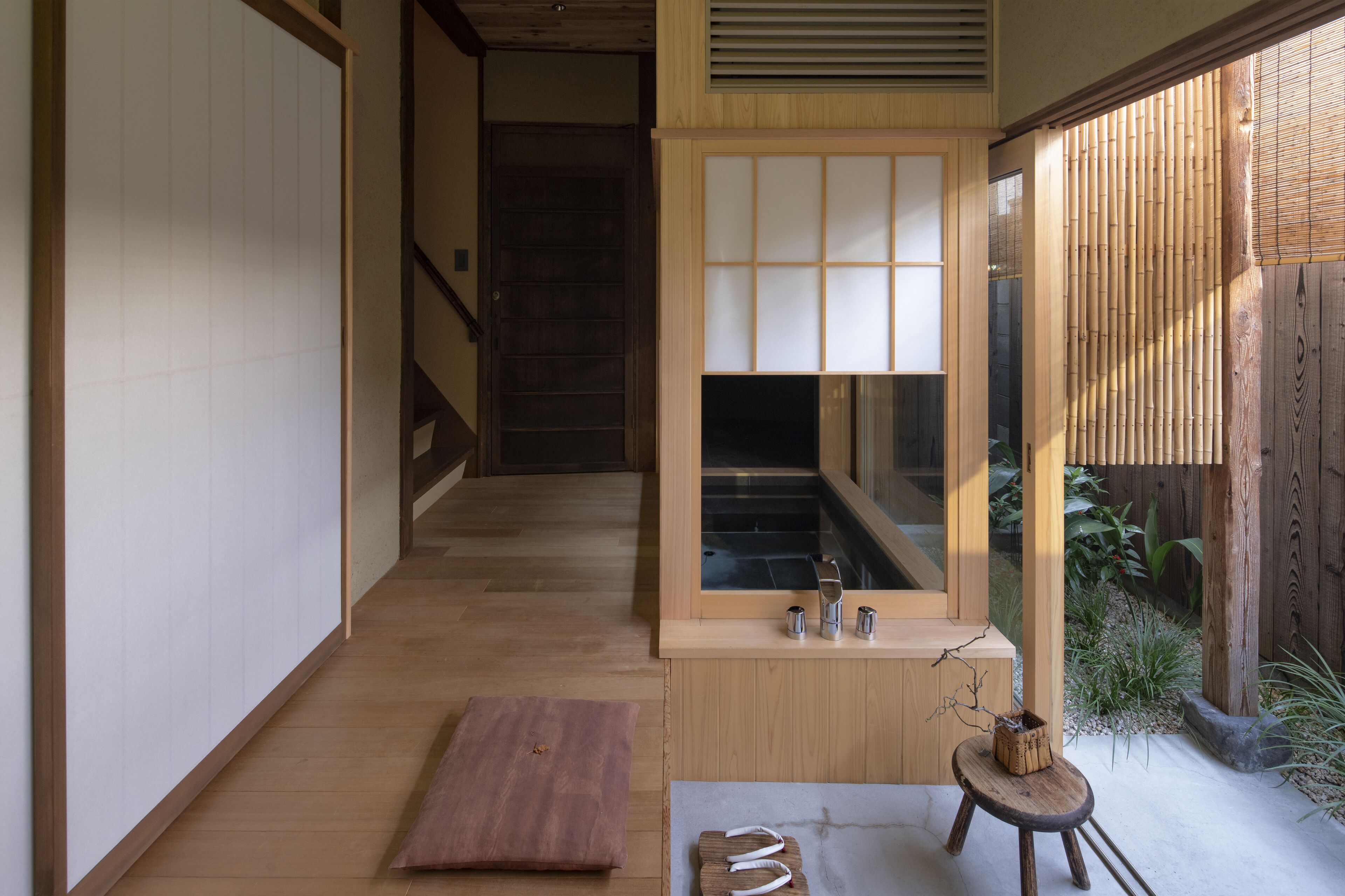 A traditional Japanese interior with a wooden floor, sliding doors, a small table with a stool, and a view of a bamboo garden.
